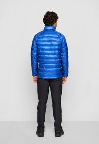 Veste puffer isolante bleue avec un col montant, présentant des surpiqûres horizontales et une finition brillante. Portée par-dessus un pantalon noir.