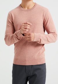 Ljusrosa stickad tröja med rund hals, ribbade ärmslut och nederkant, med en mjuk textur och figurnära design, i kombination med mörka byxor.