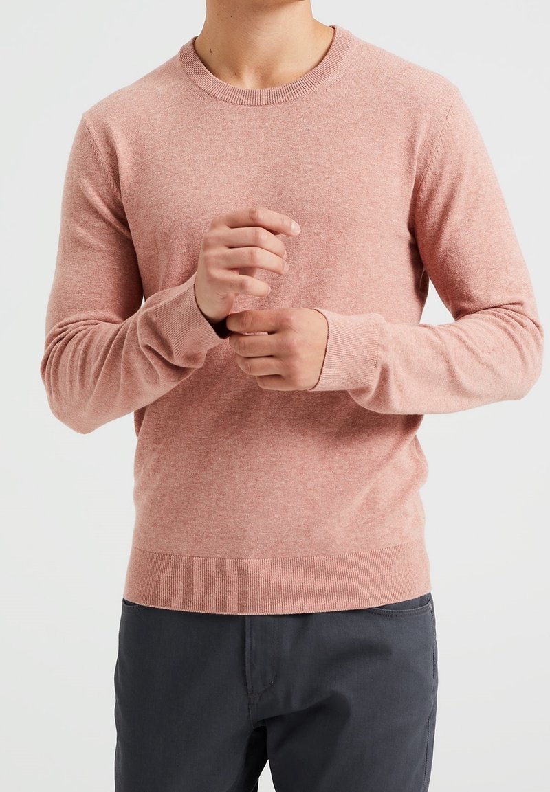 Ljusrosa stickad tröja med rund hals, ribbade ärmslut och nederkant, med en mjuk textur och figurnära design, i kombination med mörka byxor.