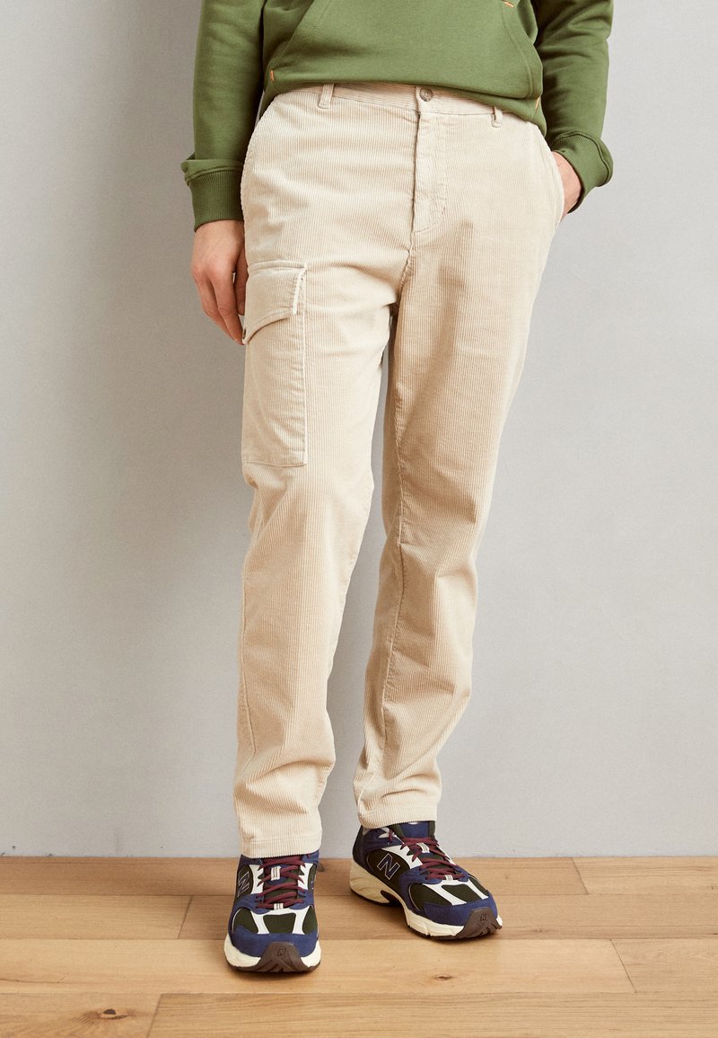 Beige kordfløyels cargo bukser med sidelommer, rett passform, og en teksturert overflate, kombinert med mørke sneakers med aksentfarger.