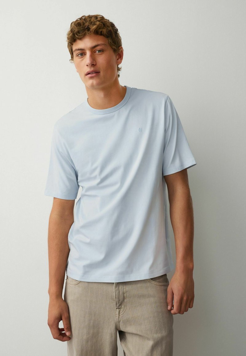 Jeune homme aux cheveux bouclés portant un T-shirt à manches courtes bleu clair et un pantalon beige, debout devant un fond blanc uni.