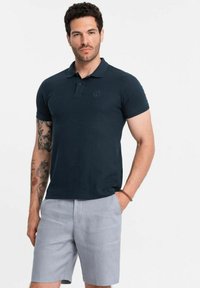 Mörkblå pikétröja med krage, korta ärmar och broderad logotyp, i kombination med ljusgrå skräddarsydda shorts. Slät textur, avslappnad passform.