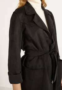 Donna che indossa una giacca in suede nera con cintura e maniche arrotolate sopra un maglione bianco a collo alto con coste.