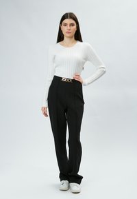 Top bianco a maniche lunghe con costine abbinato a pantaloni neri a vita alta con una cintura in catena dorata, il tutto su uno sfondo neutro.