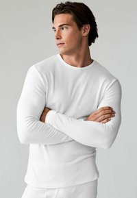 Cămașă albă cu mâneci lungi, guler rotund și textură cu nervuri, design ajustat, purtată de o persoană cu brațele încrucișate, pe un fundal neutru.