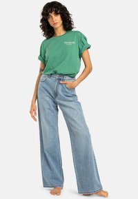 Camiseta verde de manga corta con un logo, combinada con unos pantalones vaqueros de pierna ancha color azul claro. El conjunto presenta un corte relajado y mangas enrolladas.