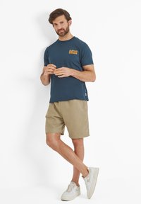 Man standing with legs crossed wearing navy blue "Endless Summer" t-shirt, beige shorts, and white sneakers against white background.