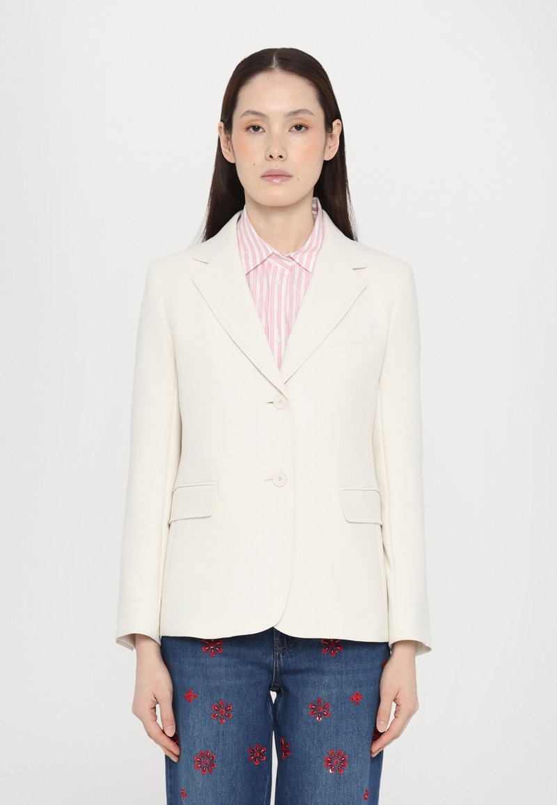 Blazer bianco su misura con due bottoni e tasche laterali, indossato sopra una camicia a righe rosa e bianche; abbinato a jeans ricamati.
