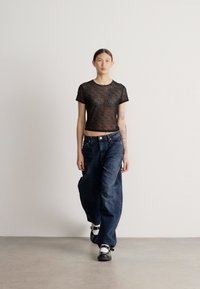 Priesvitný čierny pruhovaný krátky top v kombinácii s voľnými tmavomodrými džínsami. Modelka stojí na neutrálnej pozadí a predvádza tento outfit.