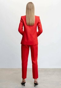 Una persona sta in piedi con le spalle alla macchina fotografica, indossando un abito rosso aderente e tacchi alti, davanti a uno sfondo bianco uniforme.