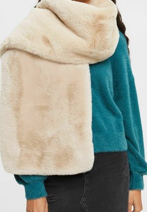 Femme portant une écharpe en fausse fourrure beige sur un pull duveteux bleu sarcelle et un jean noir déchiré, vue du cou jusqu'aux hanches.