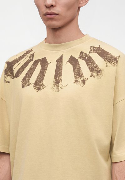 Μπεζ oversized μπλούζα με σκισμένα σκούρα καφέ γοτθικά γράμματα στο πάνω μέρος του στήθους, φορεμένη από άτομο που φαίνεται από το λαιμό και κάτω.