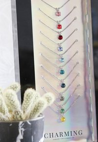 Zilveren kettingen met hangers van verschillende gekleurde edelstenen, gepresenteerd op een holografische achtergrond. Bestaat uit negen stenen in roze, rood, blauw, groen en geel.