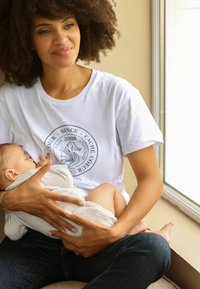 Witte t-shirt met grafisch ontwerp en tekst, gecombineerd met donkere jeans. Een persoon houdt een baby vast gewikkeld in een lichtgrijze deken.