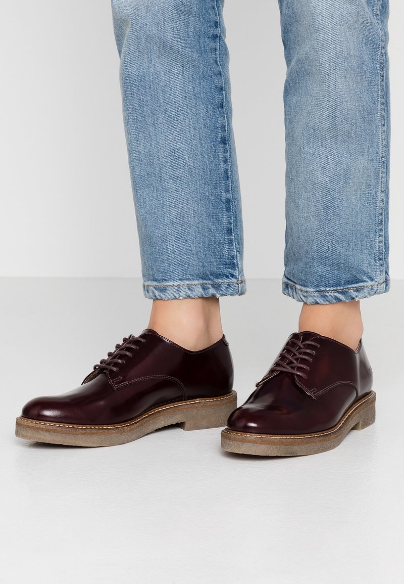Burgunderfarbene Leder-Oxford-Schuhe mit runden Zehen, Schnürung vorne und strukturierten braunen Gummisohlen, kombiniert mit hellblauen, umgeschlagenen Jeans.