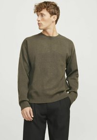Olivgrüner Strickpullover mit Rundhalsausschnitt, langen Ärmeln und gerippten Bündchen, kombiniert mit schwarzen maßgeschneiderten Hosen. Glatte Textur, lässiges Design.