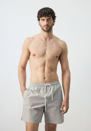 Hombre sin camiseta con cabello rizado oscuro, vistiendo pantalones cortos con cordón a rayas gris claro y blanco, de pie contra un fondo claro y liso.