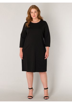 Vrouw met rood haar die een eenvoudige zwarte knie-lange jurk draagt met driekwart mouwen en zwarte enkellus hakken, neutraal staand.