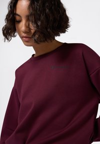 Sudadera burdeos con cuello redondo, corte holgado y logo bordado de "Champion" en verde oscuro en el pecho. Textura de tela suave.