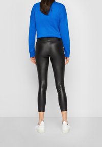 Svarta fusk läderleggings med tajt passform, tillsammans med en blå stickad tröja. Leggingsen har en slät yta och är avkortade.