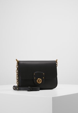 Sac bandoulière en cuir noir avec un fermoir tournant doré et une bandoulière chaîne partiellement dorée, posé sur une surface blanche devant un fond gris clair.