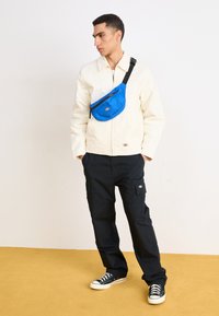 Blå midjeväska med dragkedja, bärs tvärs över kroppen över en vit jacka. Modell i svarta cargo-byxor och svarta sneakers, stående på en gul yta.