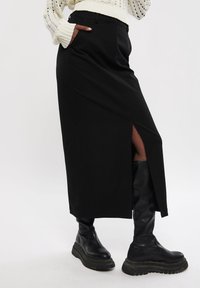 Jupe midi noire avec une fente à l'avant, des poches latérales et une texture lisse, associée à des bottes noires montantes chunky.