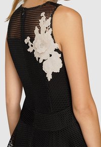 Robe sans manches en mesh noir avec une appliqué en dentelle florale blanche sur le haut du dos, portée par une personne avec des cheveux tressés.