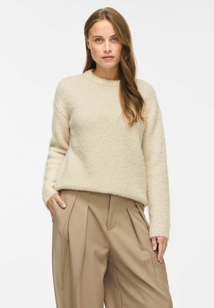 Femme aux cheveux longs portant un pull en peluche crème et un pantalon ample plissé taille haute beige, debout avec une main dans la poche.
