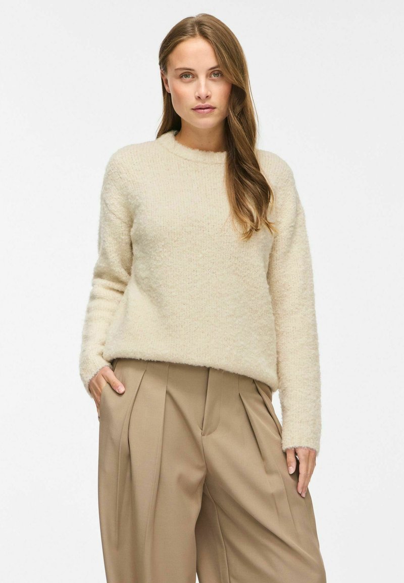 Femme aux cheveux longs portant un pull en peluche crème et un pantalon ample plissé taille haute beige, debout avec une main dans la poche.