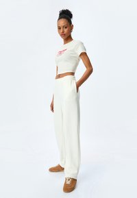 Witte cropped t-shirt met rode tekst, gecombineerd met losse witte sweatpants. Model draagt bruine sneakers met veters en een zachte textuur.