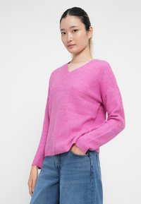 Suéter de malha em rosa brilhante com decote em V e textura canelada, com mangas longas e um ajuste descontraído, combinado com jeans azul claro.