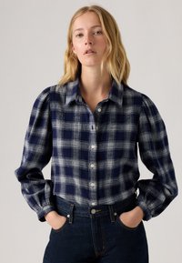 Camisa de botão xadrez azul-marinho e cinza com gola, mangas bufantes e textura de tecido macio, combinada com jeans escuros de cintura alta.