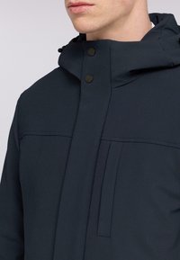 Chaqueta navy con un cuello alto, cierre de botones y costuras diagonales con una textura suave. Incluye un bolsillo frontal y capucha ajustable.