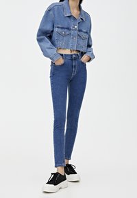 Chaqueta de mezclilla azul cortada con dos bolsillos frontales, combinada con vaqueros ajustados de talle alto azul y zapatos de plataforma negros con cordones.