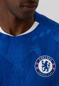 Niebieska koszulka piłkarska Chelsea Football Club z teksturowanym wzorem, białym kołnierzykiem oraz okrągłym logo z lwem i nazwą klubu.