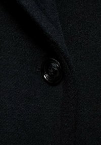 Detail van een zwarte wollen jas met een opvallende zwarte plastic knoop en een gestructureerd stofoppervlak met een zachte, donzige uitstraling.