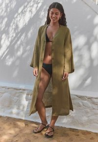 Olivově zelený lněný dlouhý kimono s knoflíkovým zapínáním, dlouhými rukávy a rozparkem, spárovaný s černým bikinim a sandály na řemínky.
