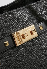 Borsa a tracolla in pelle nera con texture, dotata di un fermaglio in tonalità oro, con borchie a forma di piramide e una tracolla nera. Nome del marchio inciso sul fermaglio.