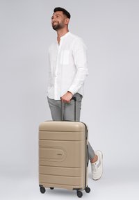 Hombre sonriente con camisa blanca y pantalones grises de pie junto a una maleta beige con ruedas, contra un fondo claro y sencillo.