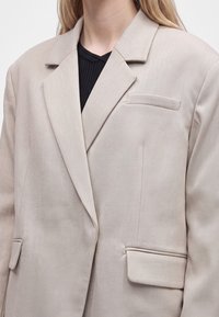 Veste blazer beige portée sur un top noir côtelé, montrant les revers, la poche poitrine et les poches à rabat, recadrée du cou jusqu'au milieu du torse.