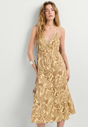 Femme aux longs cheveux ondulés portant une robe beige sans manches avec des motifs de feuilles marron et un bracelet manchette argenté à son bras gauche.