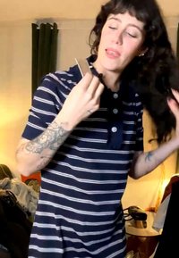 Kvinne med tatoveringer iført en stripete marineblå skjorte grer det lange, mørke håret sitt i et rotete rom med varmt lys.