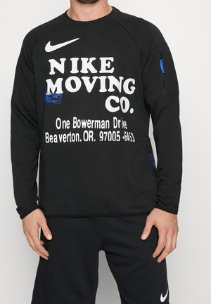 Homme portant un t-shirt noir à manches longues Nike avec un texte blanc "Nike Moving Co." et une impression d'adresse, associé à un pantalon Nike noir avec un logo blanc.