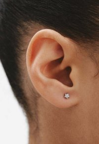 Pequeño pendiente en forma de estrella, plateado con una superficie texturizada y gemas transparentes incrustadas, llevado en el lóbulo de la oreja. Tono de piel visible en el fondo.