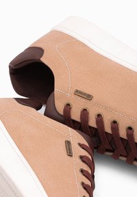 Béžové plátěné tenisky s tmavě hnědými akcenty. Mezi vlastnosti patří zaoblená špička, bílá gumová podrážka a hnědé tkaničky. Viditelný malý štítek značky.