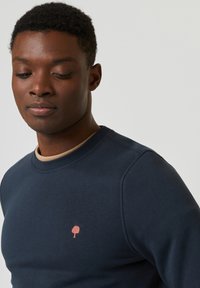 Felpa navy con collo rotondo e polsini a coste, caratterizzata da un piccolo emblema di un albero rosa sul petto. Texture liscia con un design aderente.