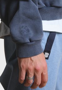 Námořnická modrá mikina s žebrovanými manžetami, na níž je malý embosovaný logo slona. Ruka v popředí, na které je stříbrný prsten, vedle světlých džínů.