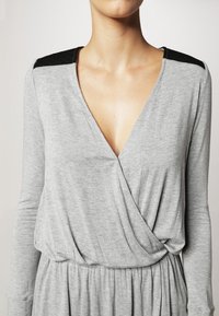 Robe grise à manches longues avec un design croisé, présentée avec des accents matelassés sombres sur les épaules, une texture lisse et une taille froncée.
