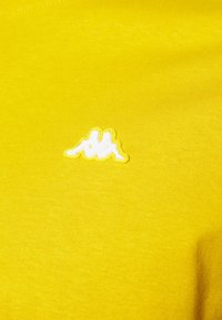 Camiseta de algodón amarilla con un pequeño logotipo bordado en blanco en el lado izquierdo. Textura suave con un acabado liso.
